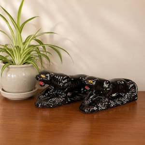 May include: Two black ceramic panther figurines with yellow eyes and red mouths. The panthers are lying down on a textured base. A potted plant with green leaves is in the background.