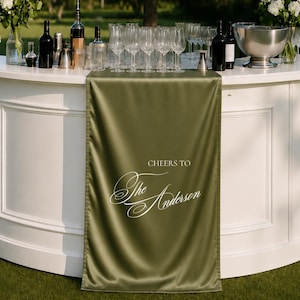 Può includere: Un runner da tavola in tessuto verde oliva drappeggiato su un bancone bianco curvo. Il runner presenta una scritta bianca stampata che recita CHEERS TO The Anderson in un mix di caratteri serif e corsivi. Il bancone è allestito con calici, bottiglie e fiori.