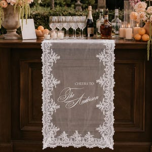 Könnte beinhalten: Ein weißer Spitzen-Tischläufer mit komplizierten Blumenmustern an den Rändern und dem mittig aufgedruckten Text "CHEERS TO The Anderson". Der Läufer liegt auf einer dunklen Holzbar und besteht aus einem transparenten Stoff mit gewelltem Saum.