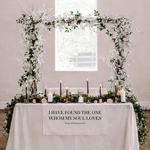 May include: A wedding reception table decorated with white flowers, greenery, and candles. The table is draped with a white cloth, and a banner displays the text "I HAVE FOUND THE ONE WHOM MY SOUL LOVES." An arch of flowers frames the table.