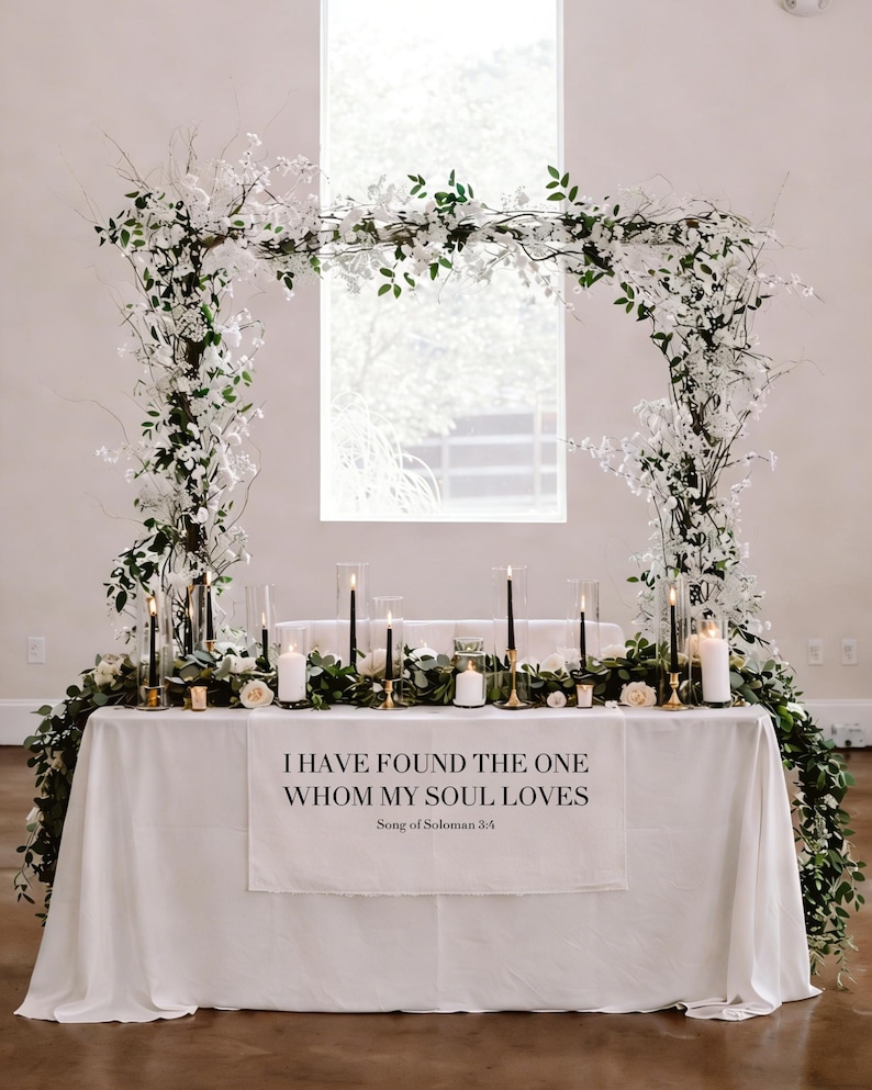 May include: A wedding reception table decorated with white flowers, greenery, and candles. The table is draped with a white cloth, and a banner displays the text "I HAVE FOUND THE ONE WHOM MY SOUL LOVES." An arch of flowers frames the table.