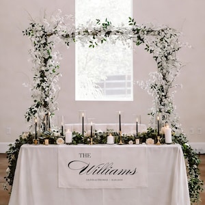 May include: A wedding reception table is decorated with a white tablecloth and floral arrangements. The table is adorned with candles, greenery, and a banner that reads "The Williams" with the date. An archway of flowers and greenery frames the table.