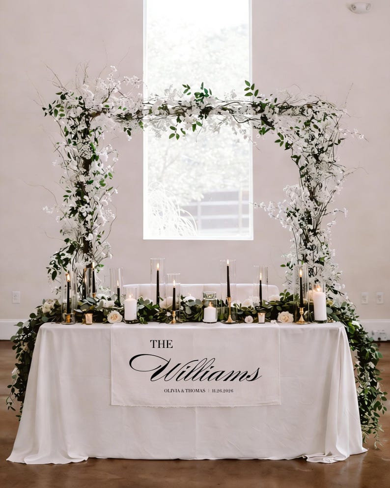 May include: A wedding reception table is decorated with a white tablecloth and floral arrangements. The table is adorned with candles, greenery, and a banner that reads "The Williams" with the date. An archway of flowers and greenery frames the table.