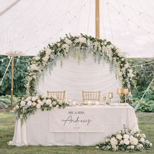 May include: A beautifully decorated wedding reception table with a white tablecloth, adorned with floral arrangements of white and blue flowers. The table features a banner with the names "Mr. & Mrs. Andrews" and the date.