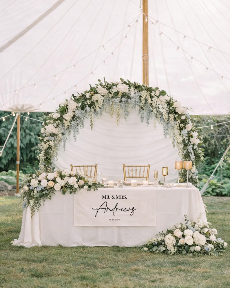 May include: A beautifully decorated wedding reception table with a white tablecloth, adorned with floral arrangements of white and blue flowers. The table features a banner with the names "Mr. & Mrs. Andrews" and the date.