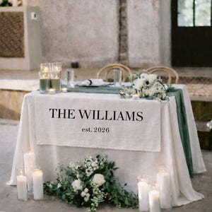 Op de afbeelding: Een bruilofttafel is gedekt met een wit linnen tafelkleed en een groene loper. De tekst "THE WILLIAMS est. 2026" staat op de voorkant van de tafel. Witte kaarsen en bloemstukken versieren de tafel.