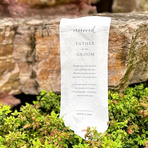 Puede incluir: Un letrero de tela blanca con las palabras "reserved" y "FATHER of the GROOM" en escritura elegante. El letrero incluye un poema y el nombre "PETER A ZEPF" en la parte inferior. El letrero se muestra al aire libre.