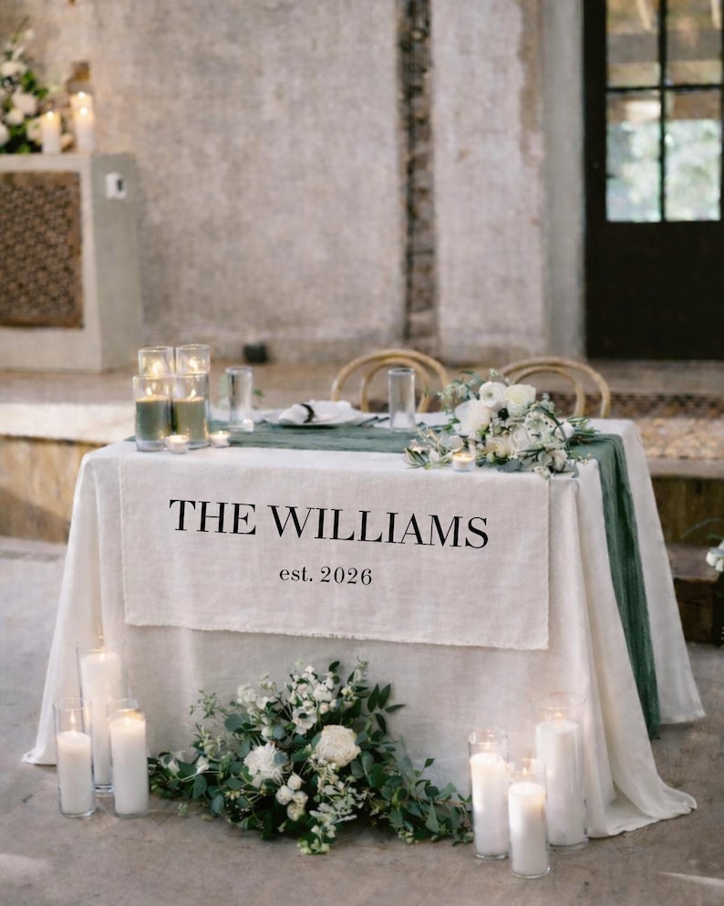 May include: A wedding reception table is set with a white linen tablecloth and a green runner. The table is decorated with white flowers, candles, and a banner that reads "THE WILLIAMS est. 2026".