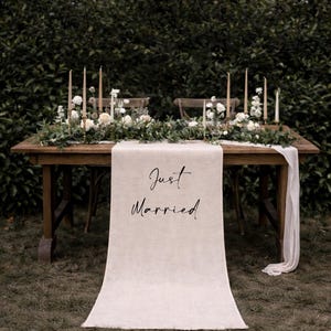 May include: A wooden table set for a wedding, adorned with floral arrangements, candles, and a linen table runner. The runner displays the text "Just Married" in elegant script, set against a backdrop of greenery.