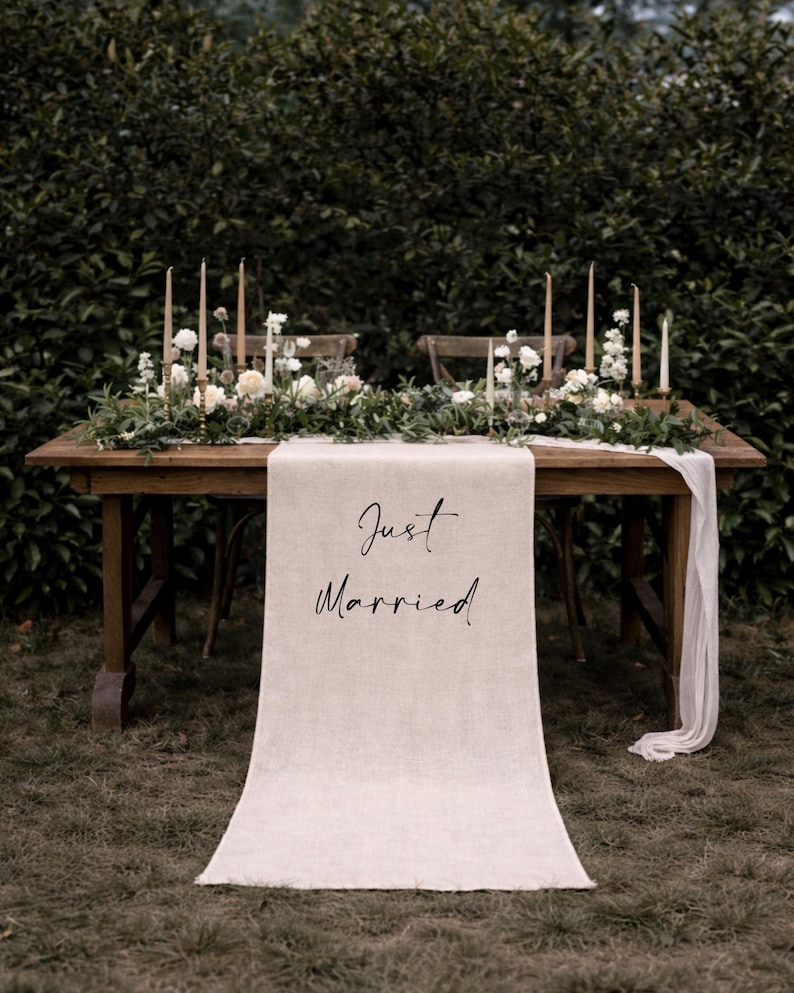 May include: A wooden table set for a wedding, adorned with floral arrangements, candles, and a linen table runner. The runner displays the text "Just Married" in elegant script, set against a backdrop of greenery.