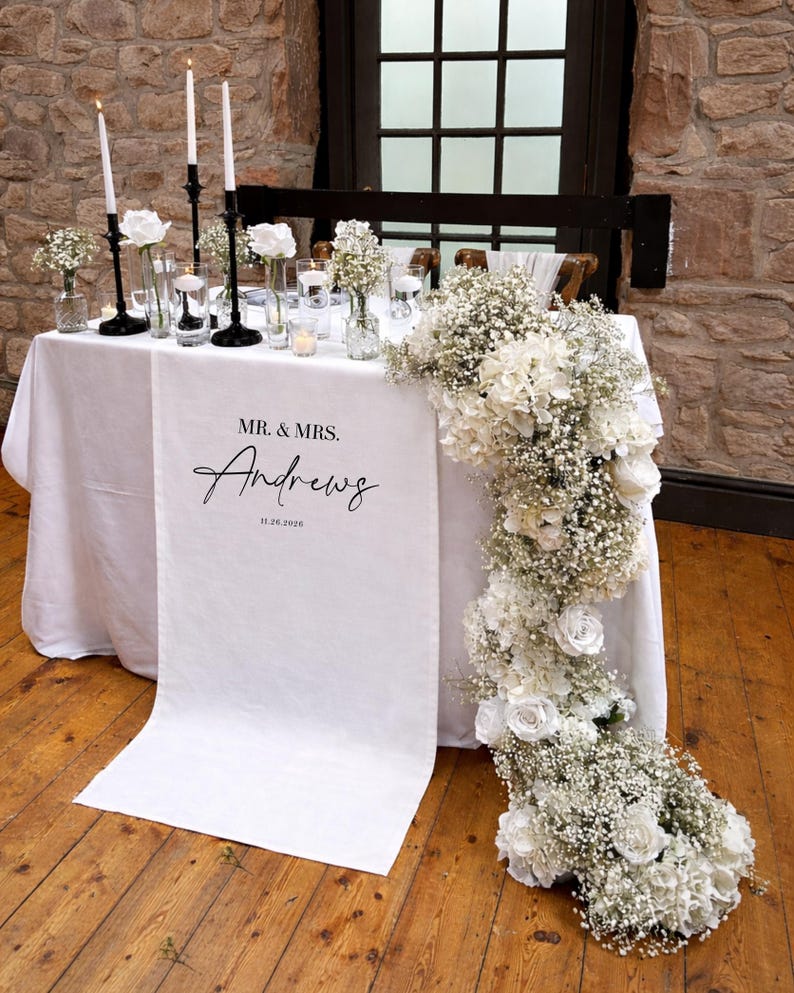 May include: A wedding reception table is set with a white tablecloth, adorned with white flowers, candles, and a personalized runner. The runner reads "Mr. & Mrs. Andrews" with the wedding date. Black candle holders and glass vases add to the elegant decor.