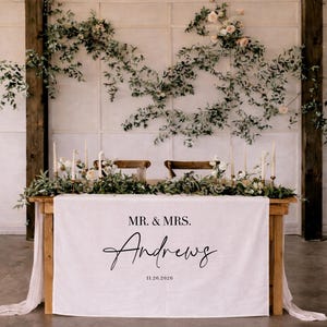Könnte beinhalten: Ein Holztisch für eine Hochzeitsfeier, bedeckt mit einer weißen Leinentischdecke. Auf der Tischdecke steht "MR. & MRS. Andrews" und das Datum "11.26.2026". Der Tisch ist mit Blumen, Grün und Kerzen dekoriert.