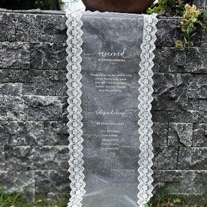 May include: A memorial sign with the text "reserved in loving memory" and a list of names. The sign is a sheer, grey fabric with white lace trim. The sign is placed on a stone wall.
