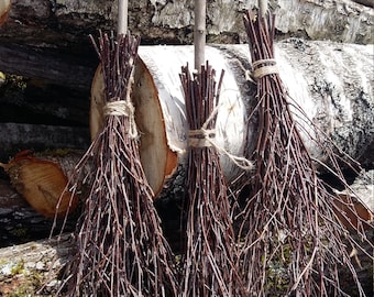 Puede incluir: Tres escobas de rama de árbol marrón con ataduras de cuerda natural, apoyadas contra una pila de troncos de abedul.