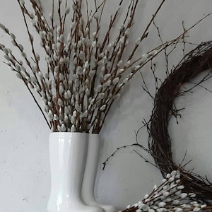 May include: A pair of white ceramic boots filled with pussy willow branches. A smaller bouquet of pussy willow branches sits on a wooden surface.