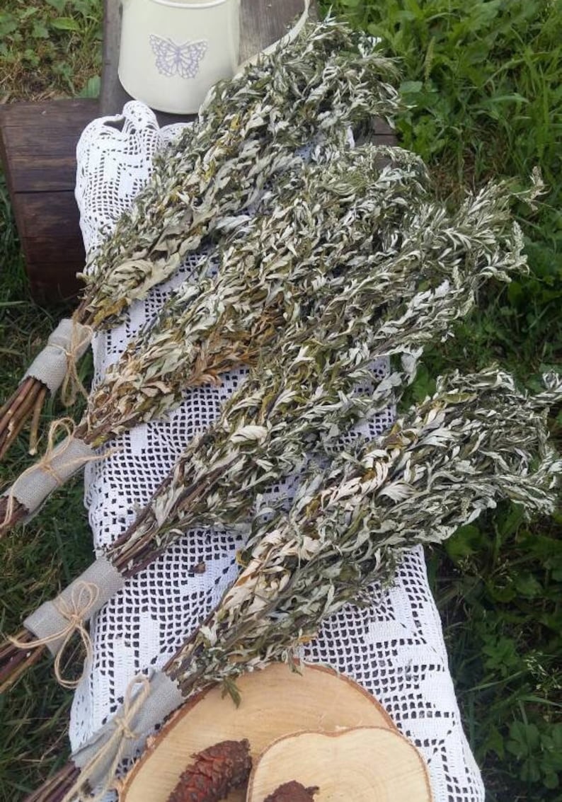 May include: Bundles of dried Artemisia, with silvery-green leaves, are tied with twine and burlap. The bundles rest on a white lace doily, with a wooden slice and a small white pitcher with a butterfly design in the background.
