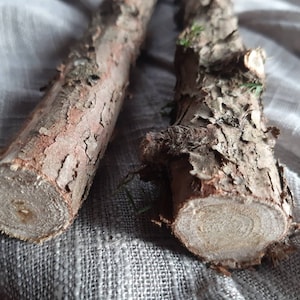 Puede incluir: Dos palos de madera marrón con corteza rugosa y un aspecto natural y rústico. Los palos están sobre una superficie de tela blanca.