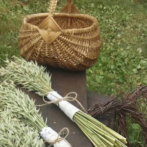 May include: A brown wicker basket with a handle is on a wooden surface, alongside bundles of dried wheat tied with white lace and twine. A wreath made of brown twigs is also present.