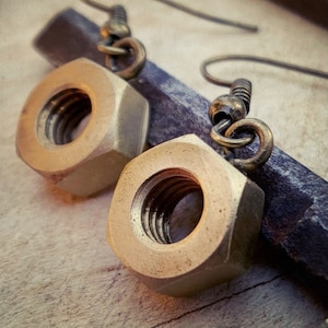 Peut inclure: Gros plan d'une paire de boucles d'oreilles en forme d'écrous de couleur laiton. Les boucles d'oreilles hexagonales ont un intérieur fileté et sont suspendues à des crochets d'oreilles en bronze antique. Elles reposent sur un objet métallique sombre.