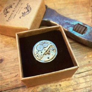 Peut inclure: Gros plan sur une bague de mouvement de montre, avec des engrenages complexes et une finition argentée. La bague est présentée dans une boîte brune avec un intérieur noir.