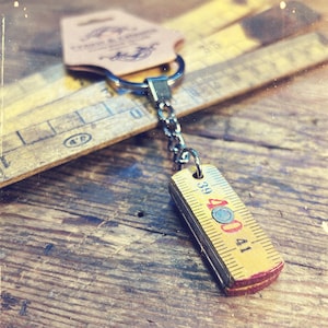 Könnte beinhalten: Ein Schlüsselanhänger im Vintage-Stil mit einem Miniatur-Zollstock. Der gelbe Zollstock hat schwarze und rote Markierungen und misst von 39 bis 41. Er hängt an einer silbernen Kette und einem Ring, mit einem braunen Anhänger. Hintergrund mit Holzlinealen.