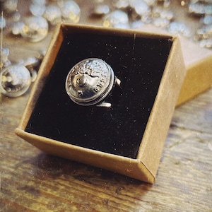 Peut inclure: Une bague en argent avec un motif circulaire détaillé, présentée dans une boîte marron avec un intérieur noir. La bague présente un motif en relief et se détache sur un arrière-plan flou avec d'autres objets argentés.