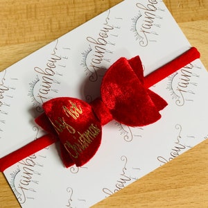 May include: Red velvet bow with a gold "My 1st Christmas" inscription on a red headband. The headband is on a white background with a repeating pattern of "Rainbow Sparkles".