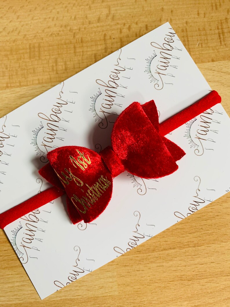 May include: Red velvet bow with a gold "My 1st Christmas" inscription on a red headband. The headband is on a white background with a repeating pattern of "Rainbow Sparkles".
