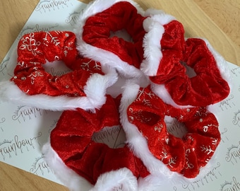 May include: A collection of four red velvet hair scrunchies with white faux fur trim. Two of the scrunchies feature a silver metallic snowflake pattern, while the other two are solid red velvet. Each scrunchie has a soft, gathered fabric texture.