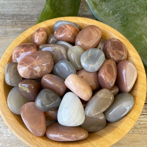 May include: A wooden bowl filled with polished moonstones in shades of gray, brown, and white. The stones have a smooth, glossy finish and vary in size and shape. The bowl is made of light-colored wood.