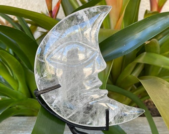 May include: A clear quartz crystal carved into the shape of a crescent moon with a face on it. The crystal is on a black metal stand.