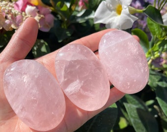 May include: Three smooth, oval-shaped rose quartz stones in various shades of pink. The stones are polished and have a translucent appearance.