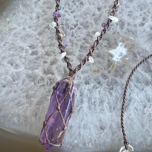 May include: A handcrafted necklace featuring a large, vibrant purple amethyst crystal pendant encased in a brown macrame net. The necklace has a brown cord with small, colorful gemstone beads.