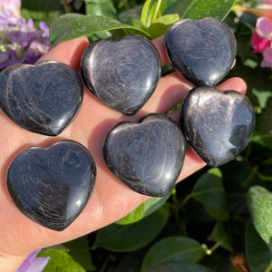 May include: Six black obsidian heart-shaped stones with silver glitter inclusions. The stones are arranged in a hand, with the tips of the hearts pointing outwards.