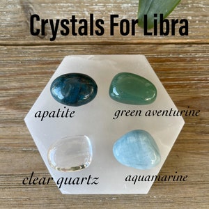 May include: Crystals for Libra: A collection of four crystals on a white hexagonal plate. The crystals are apatite, green aventurine, clear quartz, and aquamarine.