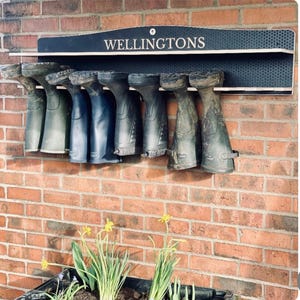 May include: A wall-mounted black boot rack with a honeycomb texture and the word WELLINGTONS printed in white. The rack holds eight pairs of rubber boots in various shades of dark green and blue, suspended upside down by their shafts.