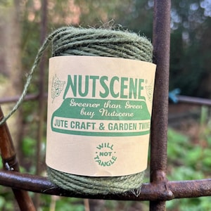 May include: A spool of olive green jute twine wrapped in a tan paper label. The label features the text NUTSCENE, Greener than Green buy Nutscene, JUTE CRAFT & GARDEN TWINE, and WILL NOT TANGLE. The twine is a natural fibre cord for crafting.