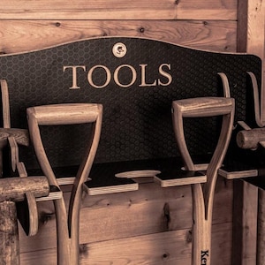 May include: A black wall-mounted wooden organiser with a honeycomb pattern and the word TOOLS printed in white. The rack features multiple cutouts and slots designed to hold various long-handled garden tools with wooden handles securely in place.