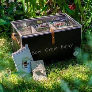 Könnte beinhalten: Eine schwarze Saatgut-Aufbewahrungsbox aus Holz mit transparentem Acryldeckel, dem Schriftzug "Sow Grow Enjoy" auf der Vorderseite und internen Trennwänden mit der Beschriftung "Herbs," "Veg," und "Flowers." Mit braunem Lederriemen.
