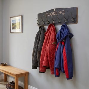 May include: A wall-mounted coat rack with a dark gray, textured rectangular backboard featuring the text "COOKE HQ" in white lettering. The rack includes seven curved, dark metal hooks, with a black quilted jacket, a red jacket, and a blue jacket.