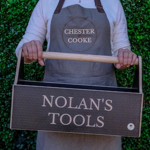 May include: A rectangular wooden tool caddy with a light-colored cylindrical handle. The front panel features a dark textured surface with the engraved text "NOLAN'S TOOLS" and a small circular logo in the bottom right corner.