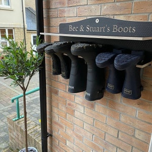 May include: A black wall-mounted boot rack with a shelf, featuring the text "Bec & Stuart's Boots" in white lettering. The rack holds six black rubber boots hanging upside down by their soles, showcasing the tread patterns against a brick wall.