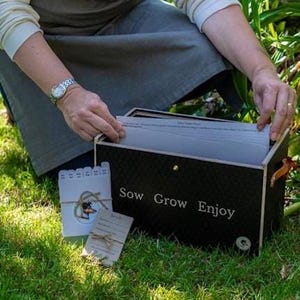 May include: A black rectangular storage box with the text Sow Grow Enjoy printed in white on the front. The box contains white paper dividers and is accompanied by a small white card with a dog illustration and a brown paper tag tied with twine.