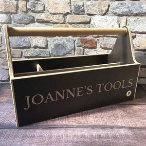 Könnte beinhalten: Ein rechteckiger Werkzeugkasten aus Holz mit schwarzer Außenseite, einem dezenten Wabenmuster und einem hellen Holzgriff. Auf der Vorderseite ist der Text JOANNE'S TOOLS in einer Serifenschrift eingraviert, zudem ist ein kleines kreisförmiges Logo zu sehen.