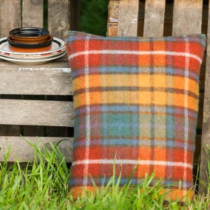 May include: A square pillow with a plaid pattern in shades of orange, red, yellow, and blue. The pillow is made of a textured fabric and is resting against a wooden crate. A stack of plates is visible in the background.