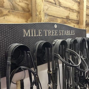 May include: A black wall-mounted tack rack with a honeycomb texture featuring the engraved text MILE TREE STABLES. The rack includes multiple curved black hooks holding several black leather horse bridles with metal buckles and rings.