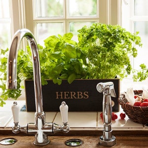 May include: A kitchen setting with a black wooden herb planter box, filled with basil and parsley. The box is labelled "HERBS" in gold. A chrome tap and sprayer are in the foreground, alongside a basket of tomatoes and garlic.