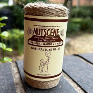 Può includere: Una bobina di spago di juta naturale avvolta in un'etichetta di carta color crema con testo marrone. L'etichetta riporta Nutscene, The Ideal Garden Twine, Natural Jute Fillis e Biodegradable, con l'illustrazione di una persona vicino a un fiore.