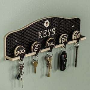 May include: A black key holder with a honeycomb pattern and the word "KEYS" in white. It has five key hooks with labels: "SHED", a flower, "DAD", a car, and a garage. Several keys hang from the hooks.