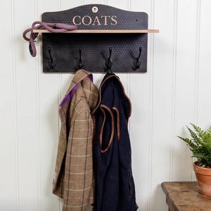 May include: A black wall-mounted coat rack with the word "COATS" in gold lettering. The rack has a shelf and four hooks, with two coats hanging. A purple leash rests on the shelf. A small fern in a terracotta pot sits to the right.